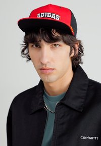 Juodas snapback kepurė su raudonu priekiniu paneliu, baltas "ADIDAS" logotipas. Modelis vilki juodą švarką, žalią marškinį, sidabrinę grandinę ir turi banguotus tamsius plaukus.