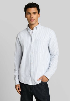 Junger Mann, der ein weißes Hemd mit dünnen senkrechten blauen Streifen und dunkle Jeans trägt, steht mit einer Hand in der Tasche vor einem schlichten Hintergrund.