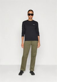 Fekete hosszú ujjú ing logóval, olívazöld nadrág és fekete cipő. A modell fekete napszemüveget visel. Egyszerű, testhez simuló kialakítás semleges színekkel.