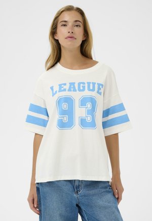 Ung kvinde iført en hvid oversize T-shirt med blå "LEAGUE 93" tekst og striber på ærmerne, kombineret med blå jeans, stående mod en hvid baggrund.