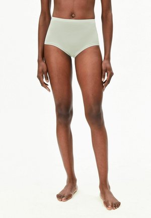 Sportliche Person steht barfuß in hochgeschnittener, hellgrüner Unterwäsche vor einfachem weißem Hintergrund.