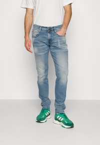 Man i ljusblå urtvättade jeans, vit t-shirt och gröna Adidas-sneakers står med ena handen i fickan mot en vit bakgrund.