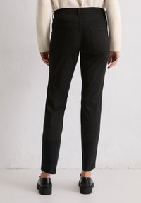 Person in schwarze Slim-Fit-Hose, schwarze Slipper und einem cremefarbenen Pullover, der vor einem hellen, strukturierten Wandhintergrund steht.