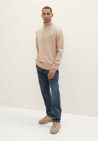 Suéter beige con cremallera y puños acanalados, combinado con jeans azul oscuro y zapatillas marrón claro. Textura suave, corte casual, diseño minimalista.