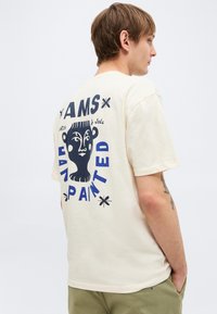 Νέος άνδρας που φοράει μια κρεμ T-shirt με μπλε γραφικό κείμενο και αφηρημένο σχέδιο προσώπου στο πίσω μέρος, στέκεται μπροστά σε απλό φόντο.