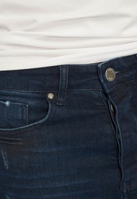 Sötétkék farmernadrág sima textúrával, fém gombos záródással, övhurkokkal, és egy kopott területtel a jobb zseb felett.