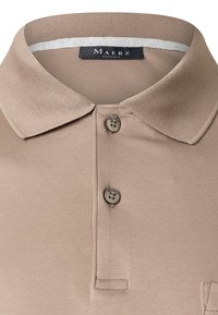 Beige Polo-Shirt mit geripptem Kragen, Drei-Knopf-Leiste und subtiler Textur. Das Markenlabel ist im Kragen sichtbar.