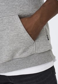 Felpa grigia con cappuccio in un tessuto testurizzato, con tasca a canguro e vita a costine, indossata sopra una camicia bianca e jeans scuri.