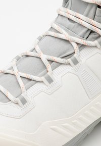 Zapatilla deportiva blanca con parte superior texturizada en tela gris y acentos. Los cordones reflectantes están entrelazados sobre la lengüeta.