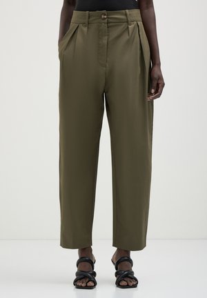 Femme portant un pantalon taillé vert olive avec des plis et des poches latérales, associé à des sandales à lanières noires à bout ouvert, se tenant sur un sol blanc.
