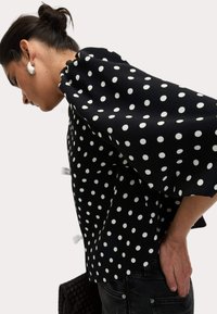 Blouse noire à pois blancs, dotée de manches larges et semi-flammées ainsi qu'un ourlet arrondi. Fabriquée dans un tissu lisse avec un léger éclat.