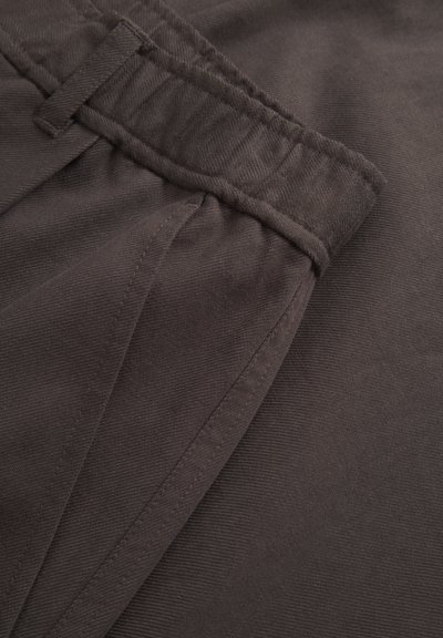 Prim-plan al unui material cafeniu închis, care arată o talie elastică și un buzunar triunghiular cusut al unei perechi de pantaloni.