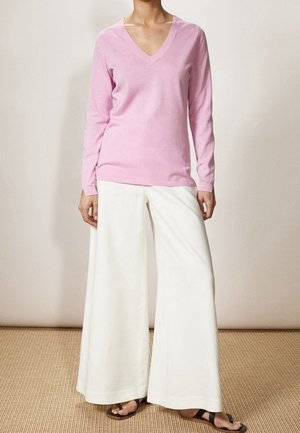Femme portant un haut rose clair à manches longues et col en V, avec un pantalon blanc à jambes larges et des sandales noires, debout sur un tapis tissé.