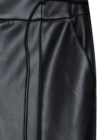 Gros plan d'un pantalon noir en simili cuir montrant le matériau texturé, la couture et une poche latérale avec de légers plis.
