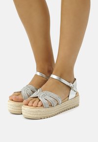 Des sandales argentées en corde avec un design tressé, dotées d'une bride de cheville ajustable et d'une semelle en jute naturel.