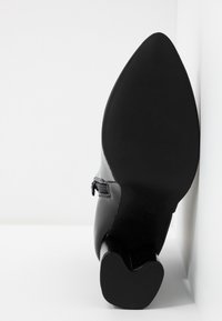 Botte en cuir noir avec un bout pointu et un talon bloc, placée debout contre un mur blanc et un sol blanc.