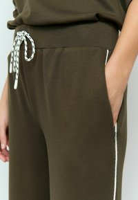 Olive green joggers with a soft texture, featuring a white drawstring and side pockets. White accents along the seams with a zip closure detail.