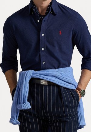Homme portant une chemise bleue marine boutonnée, un sous-vêtement à pois, un pantalon bleu marine à fines rayures, un pull bleu clair noué autour de la taille et une montre-bracelet.