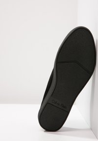 Černá podrážka bot Calvin Klein směřující ven, nakloněná proti bílému pozadí zdi a podlahy.