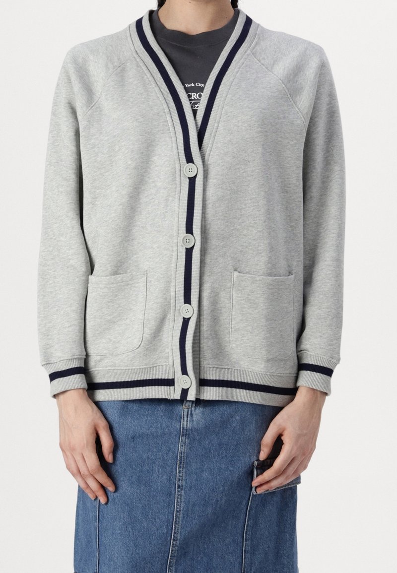 Personne portant un cardigan boutonné gris clair avec une bordure bleu marine et des poches avant, par-dessus une chemise sombre et une jupe en jean.