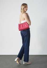 Borsa sintetica rosa con parte superiore arricciata, tracolla in catena dorata, su uno sfondo grigio. Modella indossa jeans scuri e tacchi.