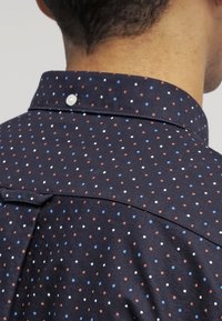 Koszula w morski wzór z małymi czerwonymi, niebieskimi i białymi kropkami, z zapięciem na guziki i teksturowaną tkaniną. Widok z tyłu podkreśla przeszycia.