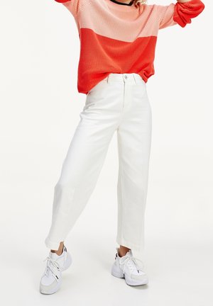 Personne portant un pull bicolore rose et rouge, un pantalon blanc taille haute et des baskets blanches sur un fond blanc uni.