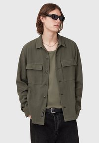 Olivengrøn, knap-oppet jakke med to brystlommer, lavet af et tekstureret stof. Båret over en matchende grøn skjorte og sorte jeans.