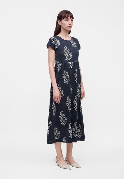 Femme debout portant une robe midi bleu foncé à motifs floraux blancs et des chaussures plates pointues beige sur un fond clair uni.