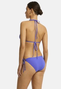 Model in een blauwe bikini, gezien van achteren, met strikjes om de hals en heupen, tegen een effen witte achtergrond.