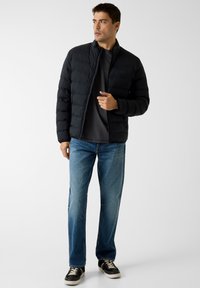 Schwarze Steppjacke mit hohem Kragen, Frontreißverschluss und gesteppter Optik, kombiniert mit blauen Jeans mit geradem Bein und schwarzen Sneakers.