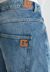 Gros plan sur un jean en denim bleu clair avec des patchs en cuir marron de la marque sur la poche arrière et la ceinture.