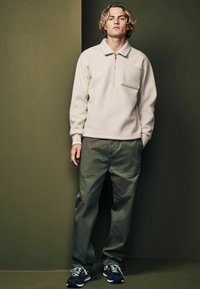 Pulover din fleece bej deschis, cu un buzunar frontal și guler cu fermoar pe jumătate, asortat cu pantaloni verzi măslinii și adidași navy pe un fundal neutru.