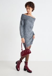 Vestido suelto gris de punto con hombros al aire, hecho de material suave, con mangas largas y cuello acanalado, combinado con botas de tobillo burdeos brillantes y un bolso.