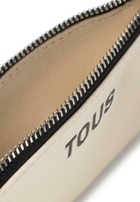 Neceser de color crema con una cremallera negra y herrajes en plata. El interior es suave con una textura sutil. "TOUS" está impreso en un lado.