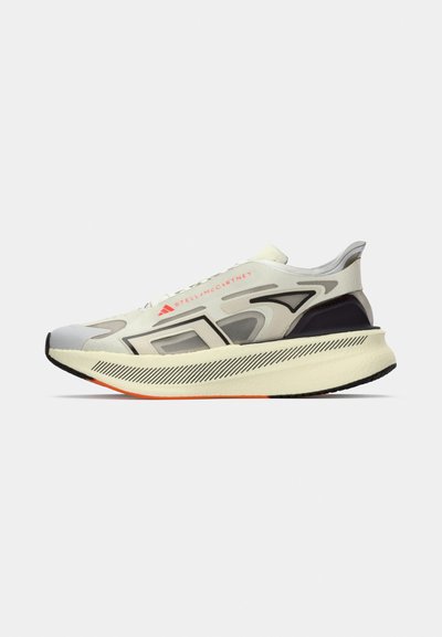 Jasno beżowe buty sportowe z kremową podeszwą, pomarańczowymi akcentami, czarnymi geometrycznymi wzorami oraz logo "Stella McCartney" z boku.