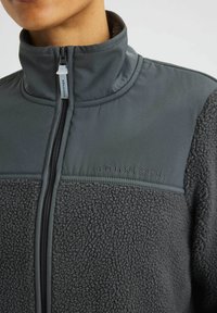 Harmaa fleece-takki, jossa on korkea kaulus, vetoketjullinen etuosuus ja teksturoitu kangas, jossa on sileä yläosa sekä brodeerattu logo.