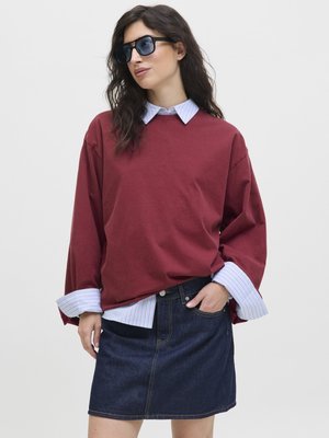 Bordeaux oversized hettegenser med rullet blå stripete mansjetter, lagd over et denimskjørt. Modell med mørke solbriller mot en nøytral bakgrunn.
