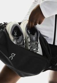 Käsin laittamassa harmaita ja valkoisia juoksukenkiä mustaan On-merkkiseen vetoketjulliseen urheilureppuun, joka on vyötäisillä.