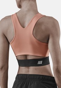 Reggiseno sportivo di colore pesca chiaro con un design a V sul retro, caratterizzato da un cinturino nero nella parte inferiore con un logo. Texture liscia, vestibilità aderente e copertura minima.