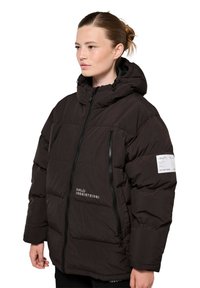 Młoda kobieta w czarnej, pikowanej kurtce z kapturem, z zamkami z przodu i napisem z logo na piersi oraz na rękawie, zwrócona w lewo na białym tle.