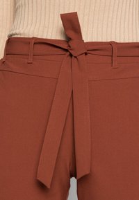 Bruine high-waisted broek met een gestrikt taille-detail. Glad materiaal en gestructureerd ontwerp met riemlussen en zijzakken.