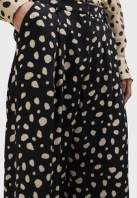 Čierne nohavice s veľkým krémovým bodkovaným vzorom, s plisovaním a bočnými vreckami, vyrobené z hladkej, ľahkej tkaniny.