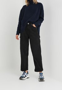 Marinblå stickad turtleneck-tröja, svarta löst sittande byxor med upplindade fållar och kraftiga blå och vita sneakers. Slät textur, avslappnad design.