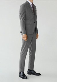 Grijs op maat gemaakt kostuum met inkepingen op de revers, twee voorkruiden, slim-fit broek en zwarte leren schoenen. Witte pochet als accent.