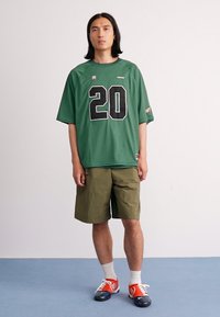 Young man with long hair wears oversized green jersey with number 20, olive cargo shorts, white socks, and red and navy sneakers.