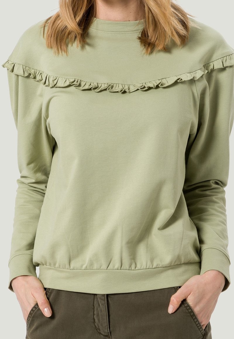 Femme portant un haut à manches longues vert clair avec un détail à volants sur la poitrine et un pantalon vert foncé, les mains dans les poches.