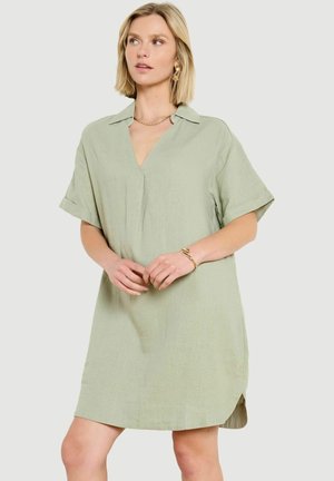 Femme portant une robe ample vert clair à manches courtes avec col et encolure en V, accessoirisée de bijoux en or, debout devant un fond uni.