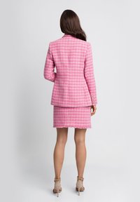 Różowa marynarka tweedowa o strukturalnym fasonie, w komplecie z dopasowaną spódnicą z postrzępionymi brzegami. Oba elementy zdobione są złotymi guzikami.