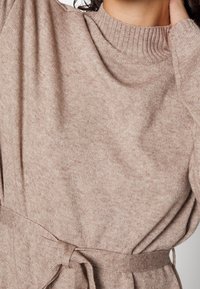 Jasno-brązowy sweter z żebrowanym wysokim dekoltem i długimi rękawami. Posiada wiązanie w talii oraz miękką, teksturowaną tkaninę.